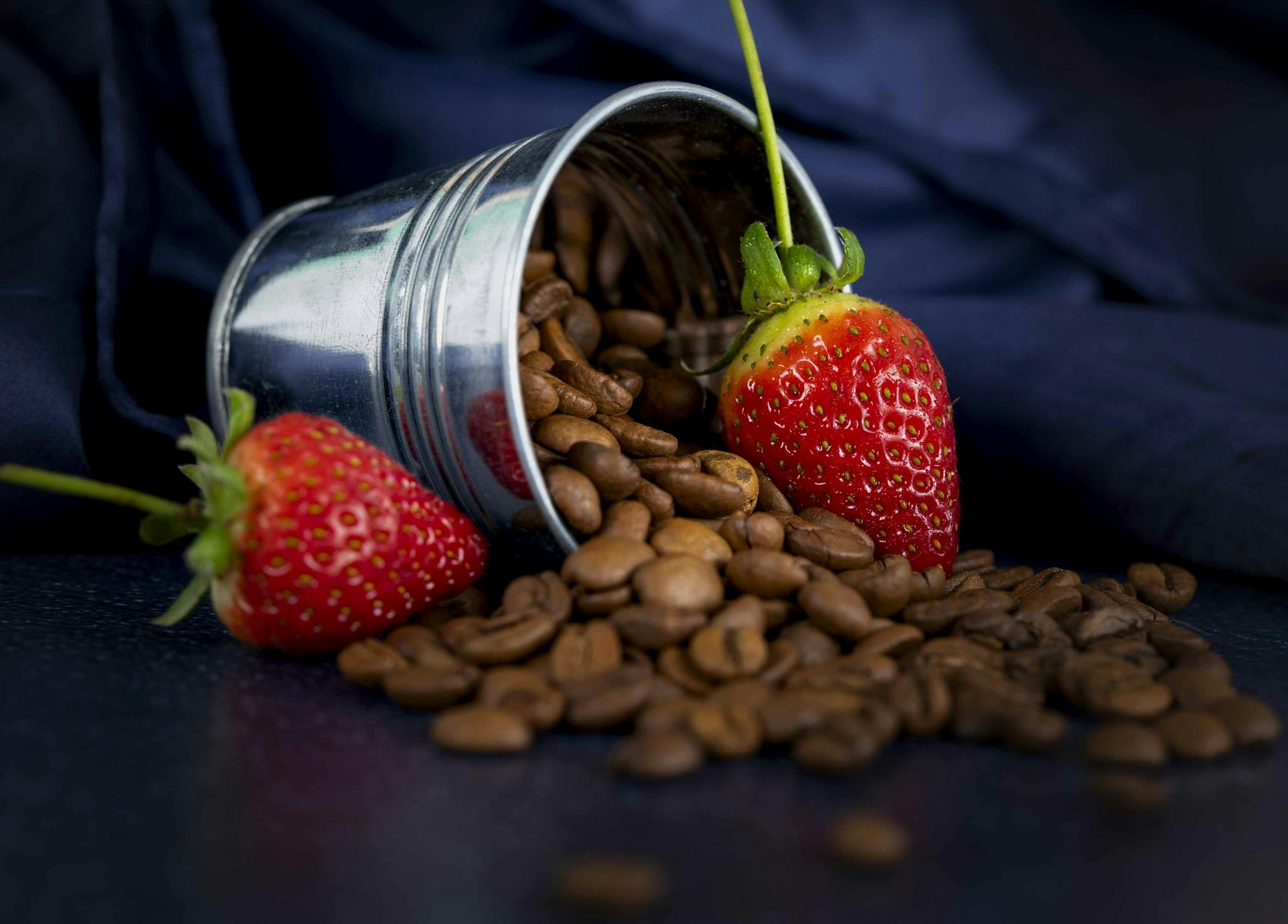 A tin can spills roasted coffee beans alongside fresh strawberries on a dark surface.