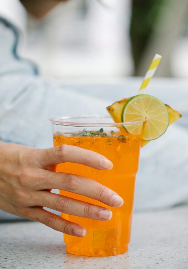 Close-up of a hand holding a citrus and pineapple juice with a straw, garnished with lime and pineapple.