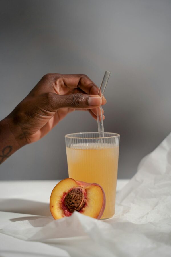 Close-up of a hand holding a glass with peach juice and straw, beside a fresh peach.