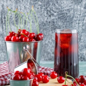 Juice with cherries, cutting board, kitchen towel in a jug on plaster and grungy background, side view.