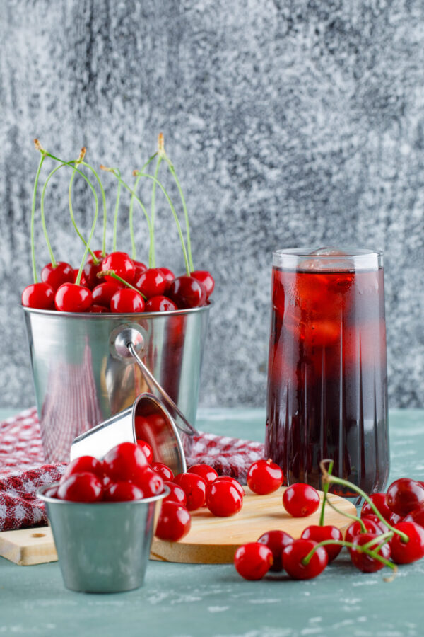 Juice with cherries, cutting board, kitchen towel in a jug on plaster and grungy background, side view.